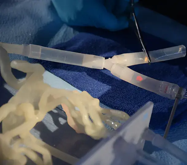 Close-up of a surgical training setup using translucent 3D-printed anatomical models and tubing, with a gloved clinician manipulating instruments on a blue drape.
