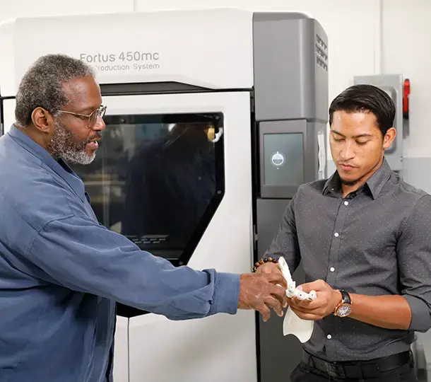 Instructor and student examining a 3D-printed prosthetic hand while standing beside a Stratasys Fortus 450mc 3D production system in a training environment.