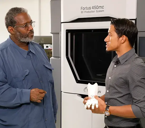 Instructor demonstrating a 3D Printed prosthetic hand to a learner while standing beside a Stratasys Fortus 450mc 3D production system.