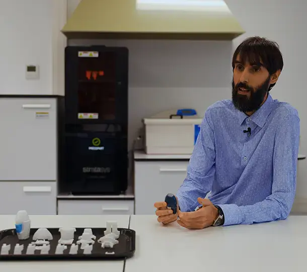 Engineer sitting in a lab beside a tray of tiered medical device prototypes, holding one prototype in his hands, with a Stratasys 3D printer and other lab equipment in the background.