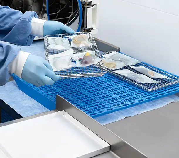 Gloved technician placing sterilized, individually wrapped 3D-printed surgical training models into wire trays on a blue sterile mat near an autoclave.