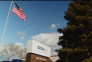 American Flag flying over a Stratays Direct sign for their additive manufacturing facility